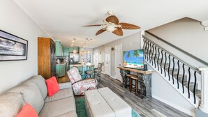 A spacious living room with a sofa, ottoman, and chairs, a kitchen area, and a staircase with a railing. - Gulf View Condo | Serene Escape with Heated Pool & Hot Tub (Corpus Christi)