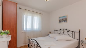 Ein Schlafzimmer mit Bett, einem Fenster mit durchsichtigen Vorhängen, einem Nachttisch mit Pflanze und einem Bild an der Wand.