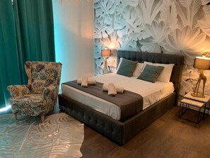 A neatly made bed with pillows and a blanket, a patterned armchair, and a decorative wall with leaf-like designs. - Suite Lame 37 Rental Rooms (Bologna)