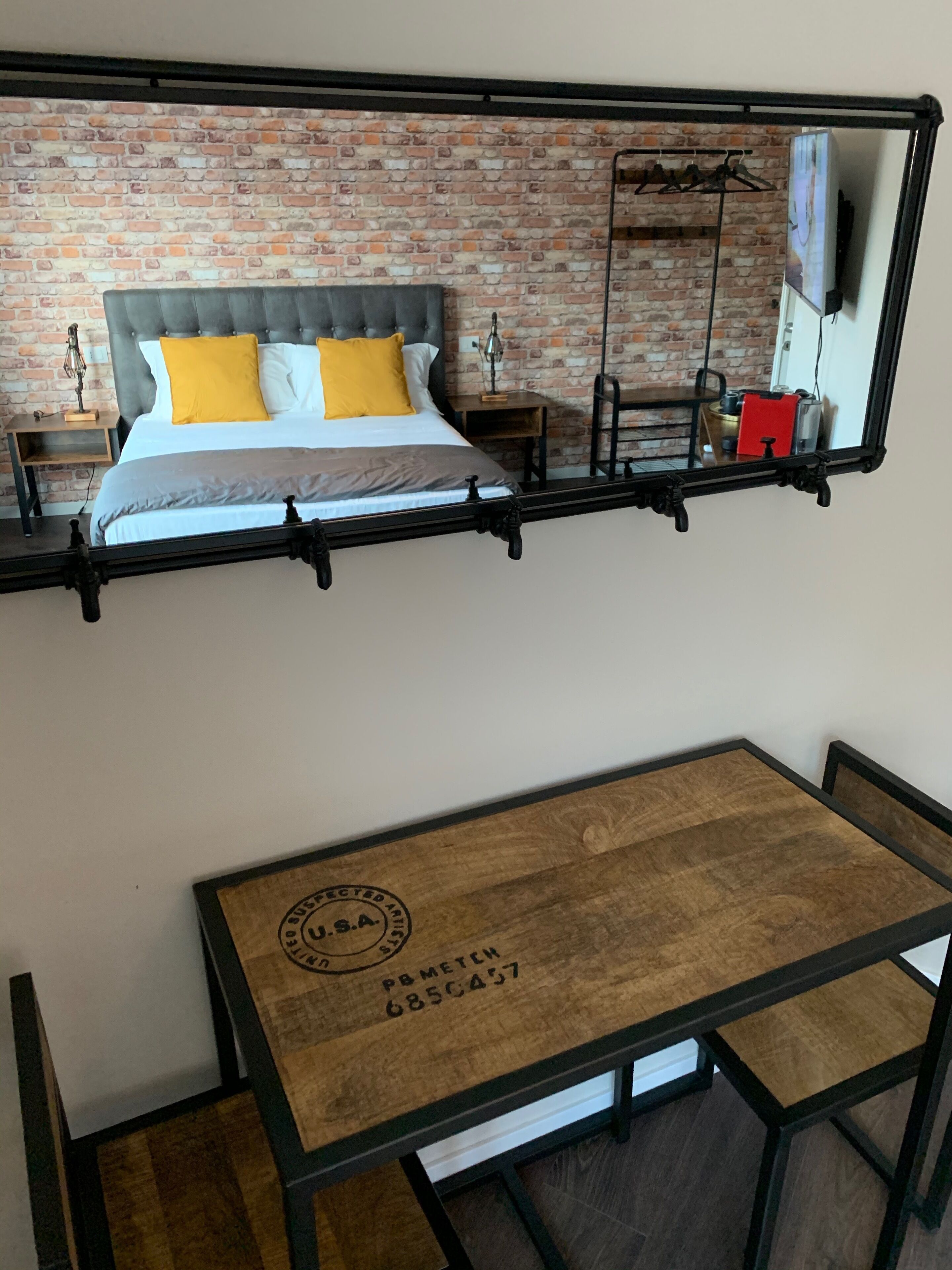 Uma cama com cabeceira estofada, uma mesa de madeira com carimbo dos EUA e uma parede de tijolos.