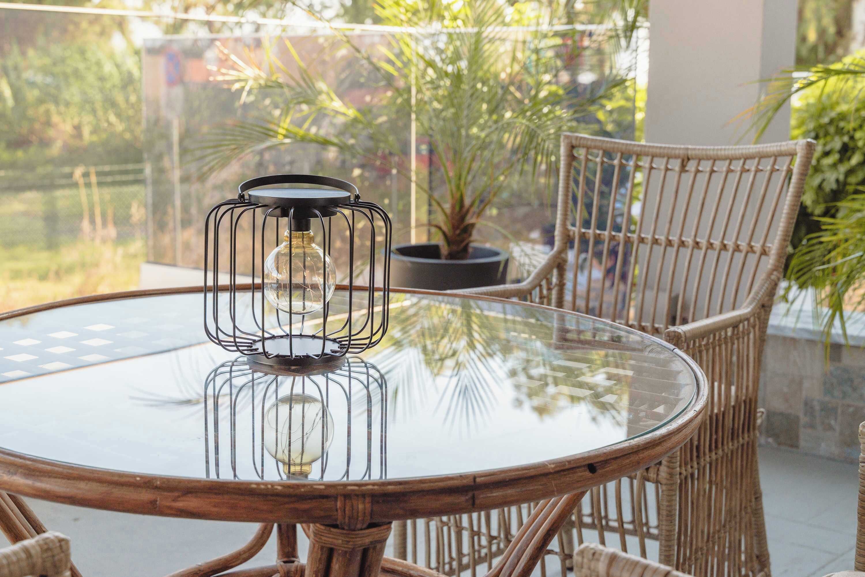 A round glass table with a metal candle holder, surrounded by wicker chairs, on a balcony with potted plants and a view of greenery.