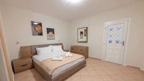 A bedroom with a bed, nightstands, a wooden dresser, a door with a window, and two wall-mounted paintings. - Seaside Nest in Kourouta Steps From the Beach (Ilida)