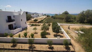 Een modern wit gebouw met een balkon, omgeven door een aangelegde tuin met bomen en een helder uitzicht op zee en bergen op de achtergrond.