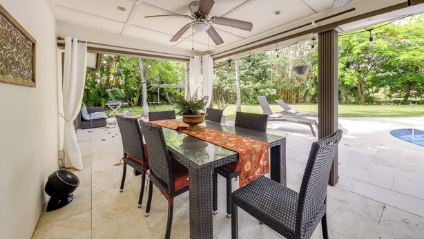An outdoor patio with a glass-top table, wicker chairs, a ceiling fan, and a view of a backyard with a pool and greenery. - Fishing Dock & Canal Views: Fort Lauderdale Home! (Fort Lauderdale)