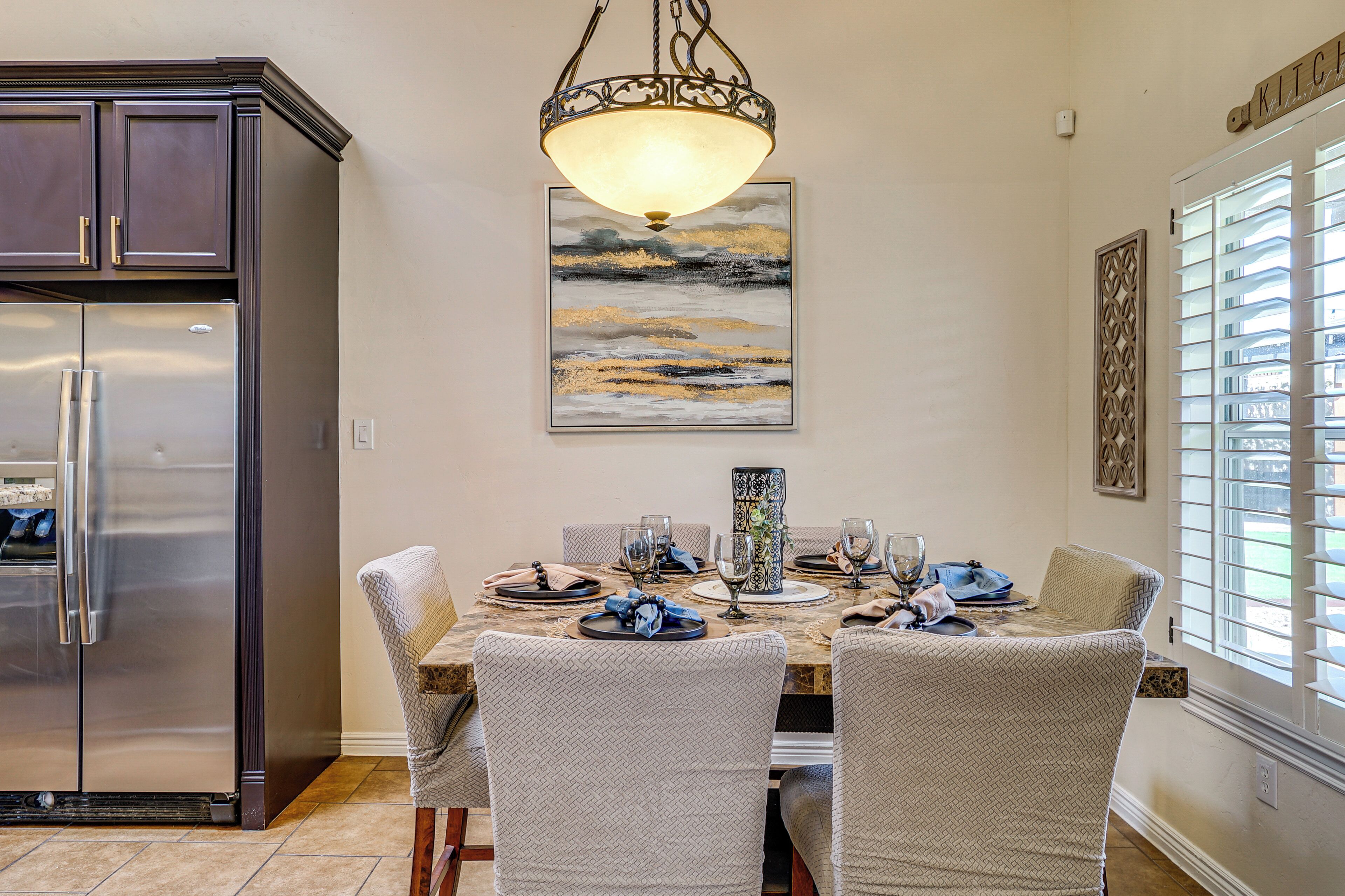 A dining area with a marble table, four upholstered chairs, a chandelier, a refrigerator, and a window with blinds.