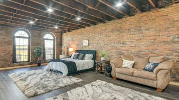 A bedroom with a brick wall, a bed with a green headboard, a beige sofa, and two large windows.