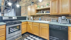 A kitchen with wooden cabinets, a stainless steel stove, a dishwasher, and a brick wall. - Deck + River View: Loft in Downtown Hannibal (Hannibal)