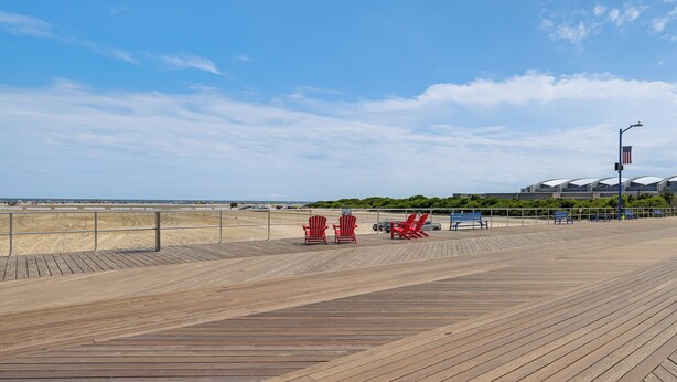 Une promenade en bois avec des chaises Adirondack rouges et bleues, une plage en arrière-plan et un bâtiment surmonté d’un dôme.