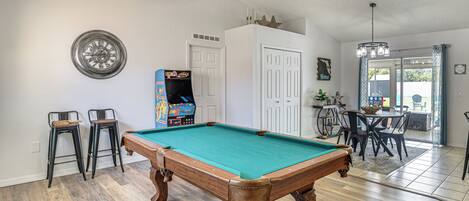 A pool table with two stools, a ceiling fan, and a door leading to a dining area.