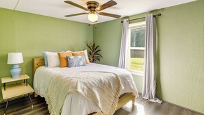 Une chambre à coucher comprenant un lit, une table de chevet, une lampe, un ventilateur de plafond et une fenêtre avec des rideaux.