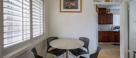 A dining area with a round table and four chairs, a painting on the wall, and a kitchen in the background.