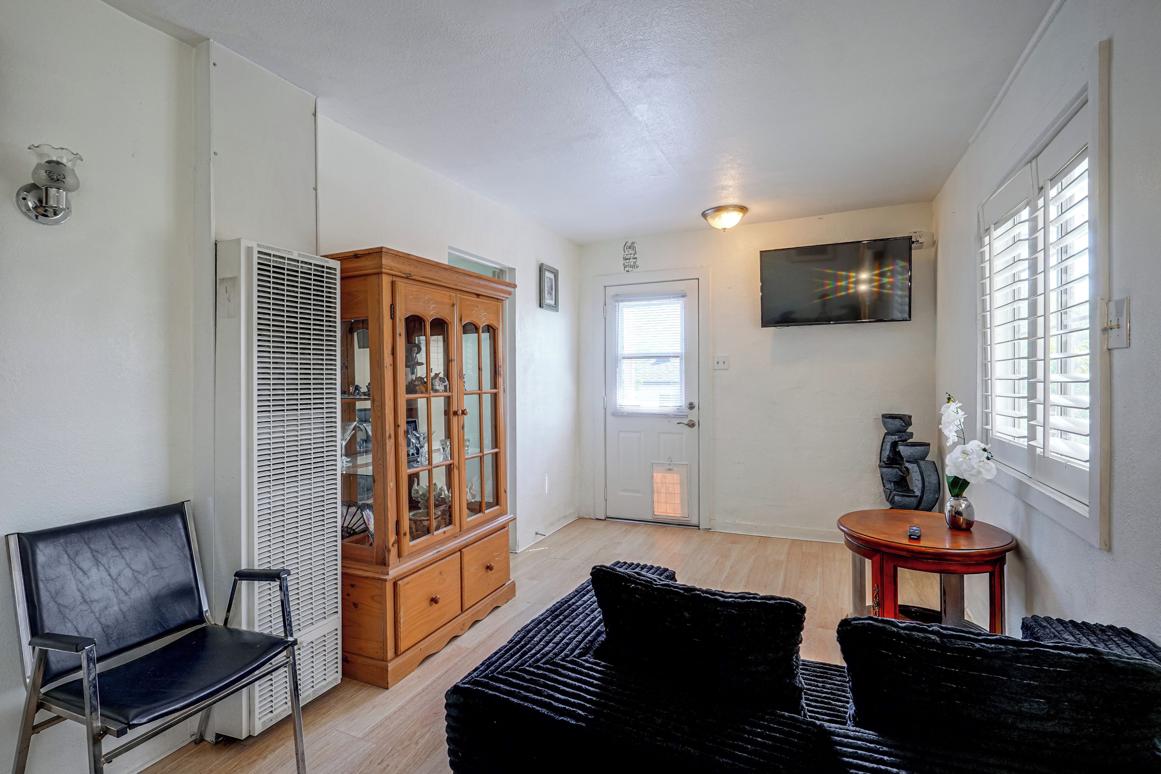 Une pièce comprenant un meuble en bois, une chaise noire, une petite table avec un vase et un téléviseur fixé au mur.