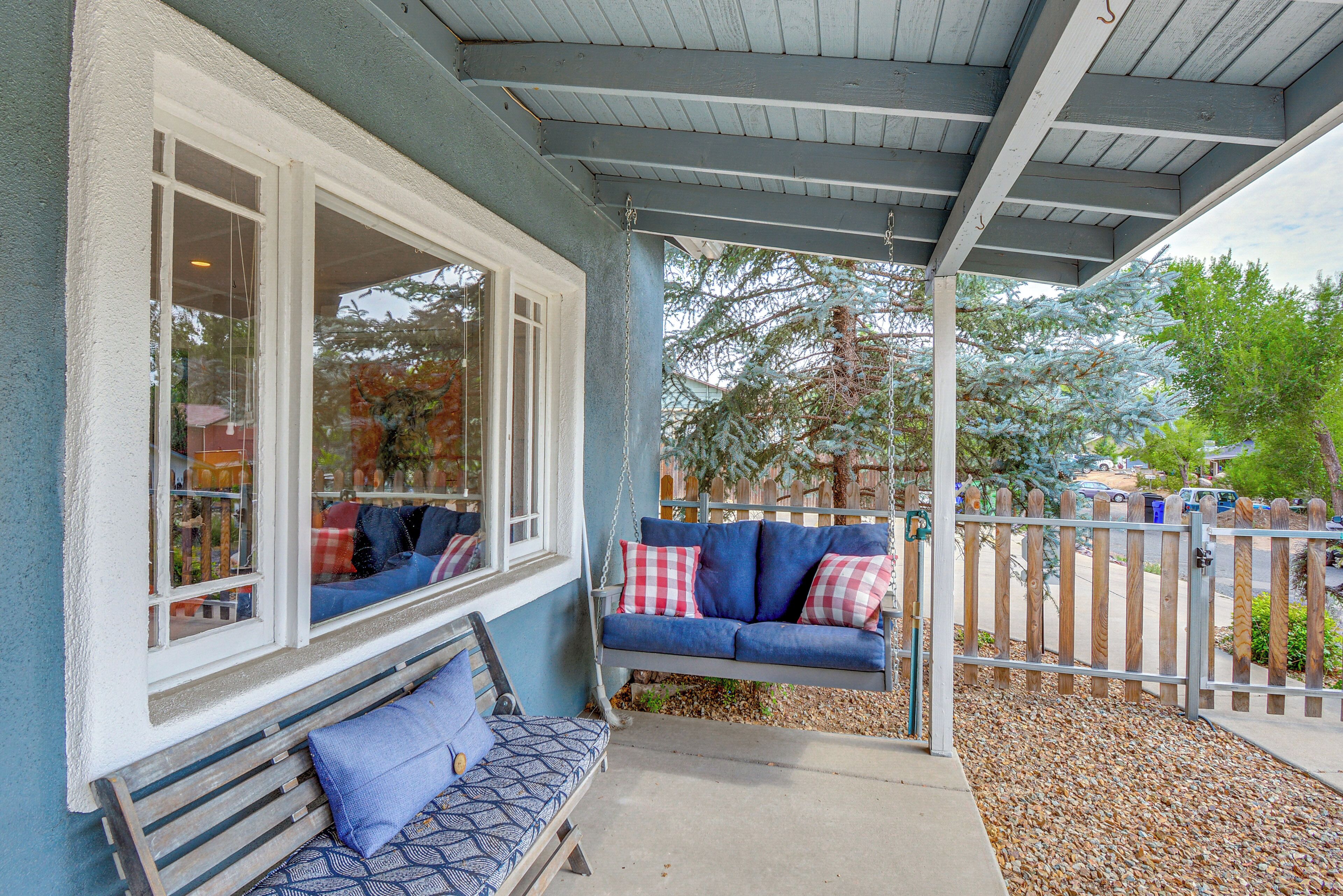 A porch with a blue bench and a swing seat, a wooden fence, and a view of trees and parked cars.
