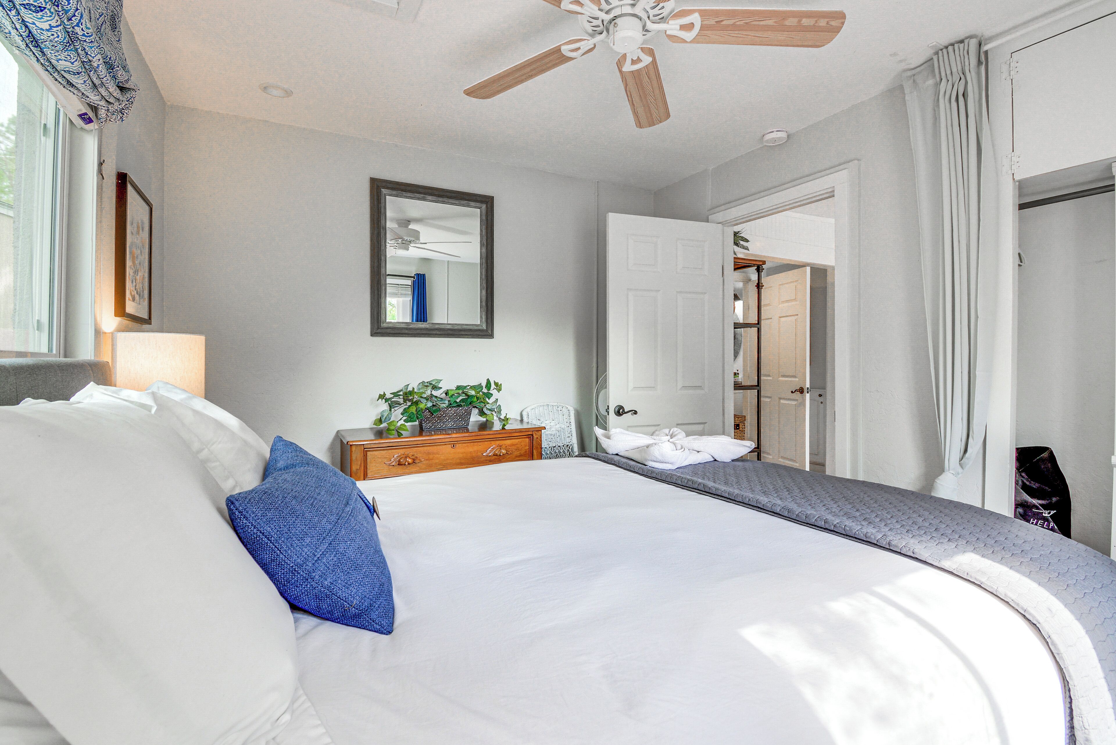 Un lit bien fait, avec des oreillers blancs et bleus, une commode en bois dotée d’un miroir et un ventilateur de plafond.