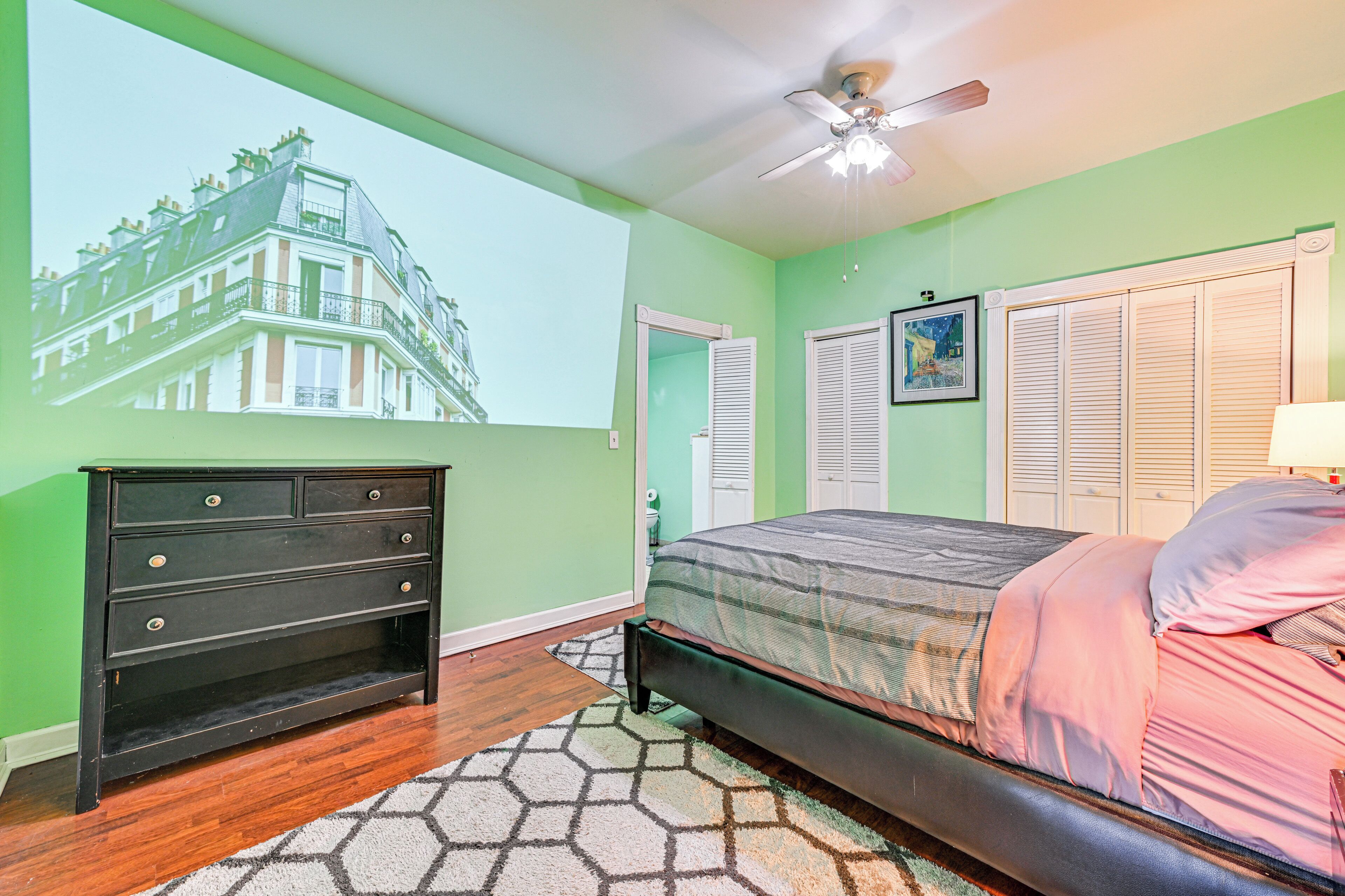 Une chambre à coucher avec un lit, une commode, un ventilateur de plafond et une vue sur un immeuble par la fenêtre.