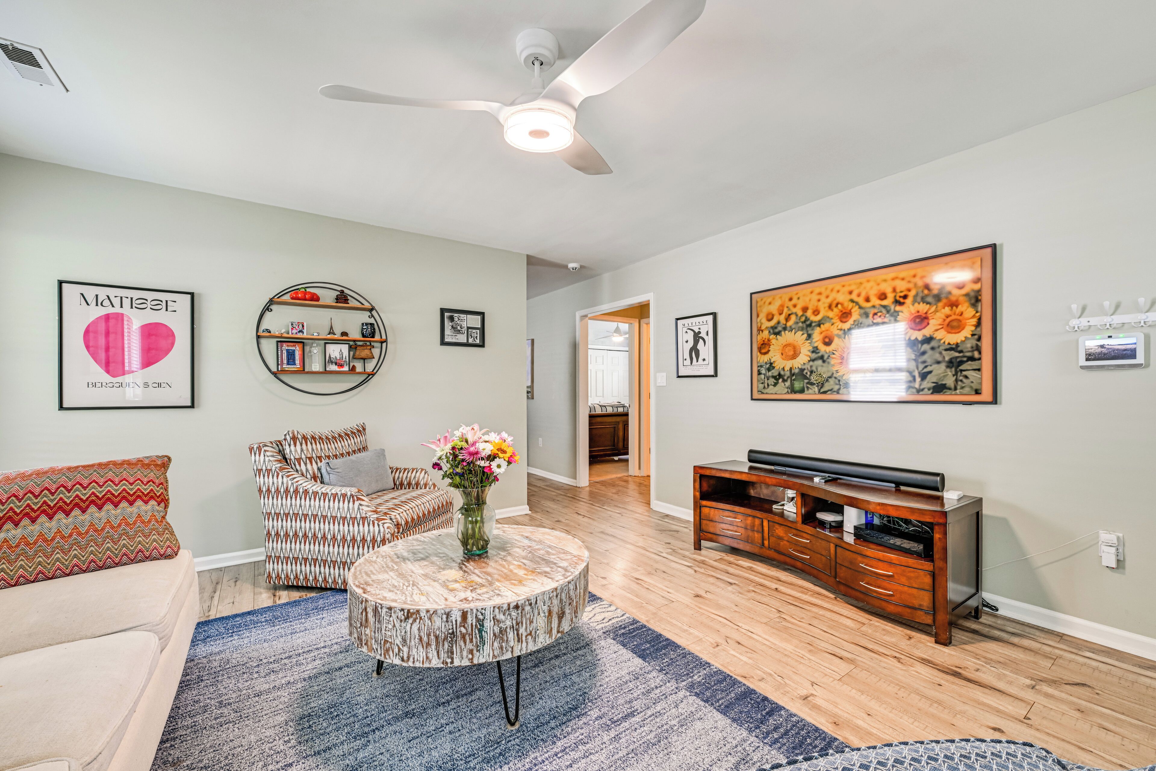 A living room with a ceiling fan, a round coffee table with a vase of flowers, a sofa, a TV stand, and a wall with framed artwork.