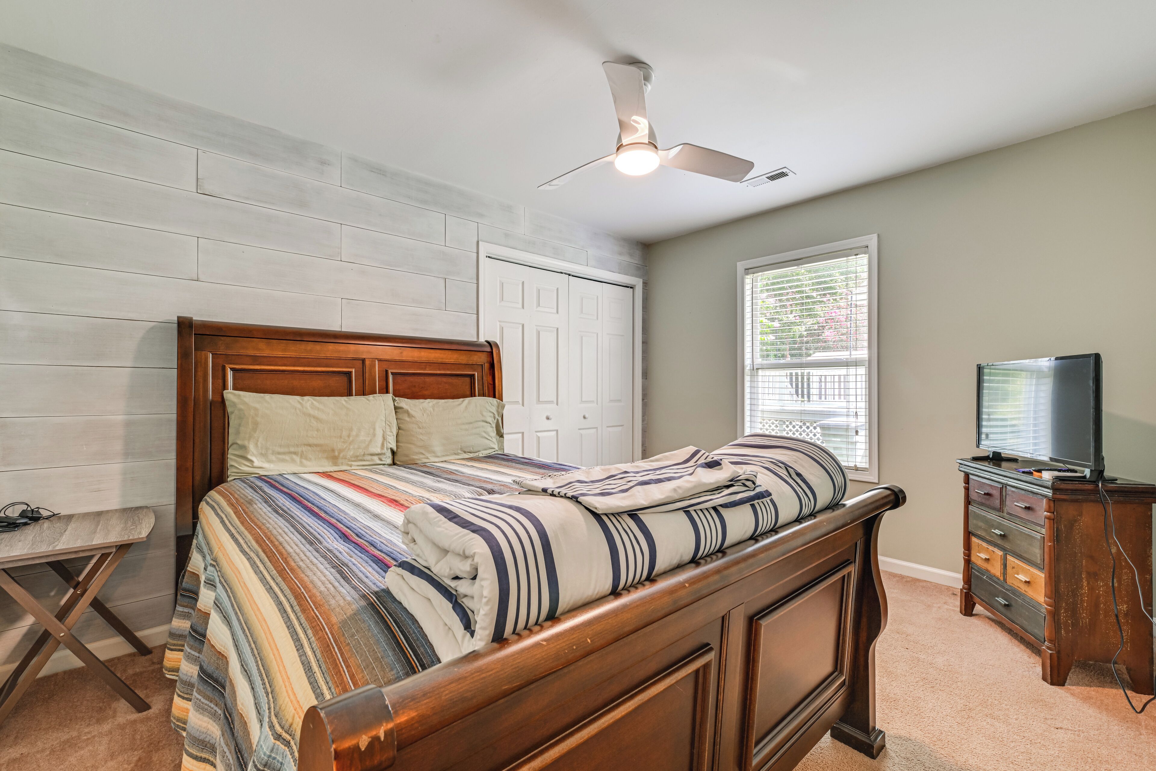 A bedroom with a wooden bed, a ceiling fan, a window with blinds, and a TV on a wooden dresser.