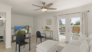 A modern living room with a flat-screen TV, a ceiling fan, a dining area with a table and chairs, and a large window with a view of the sea. - Charming Condo Across the Street from the Beach (St Simons Island)