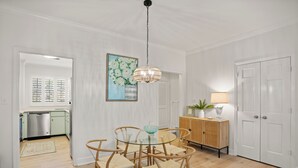 A dining area with a glass table and wicker chairs, a hanging light fixture, a painting, and a wooden cabinet with a lamp. - Charming Condo Across the Street from the Beach (St Simons Island)