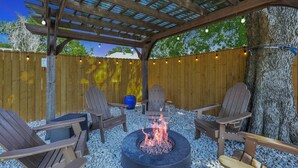 Patio esterno con un braciere, sedie di legno e un pergolato.