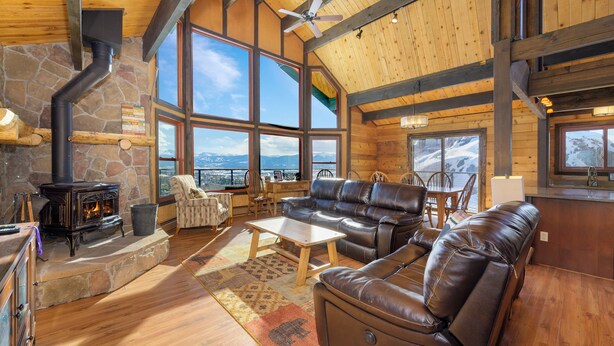 A cozy cabin interior with a stone fireplace, leather seating, large windows, and a view of the mountains. - 273 Fallen Rock BIG Views 4 Bed Home, Pet Friendly + Hot Tub! (Tabernash)