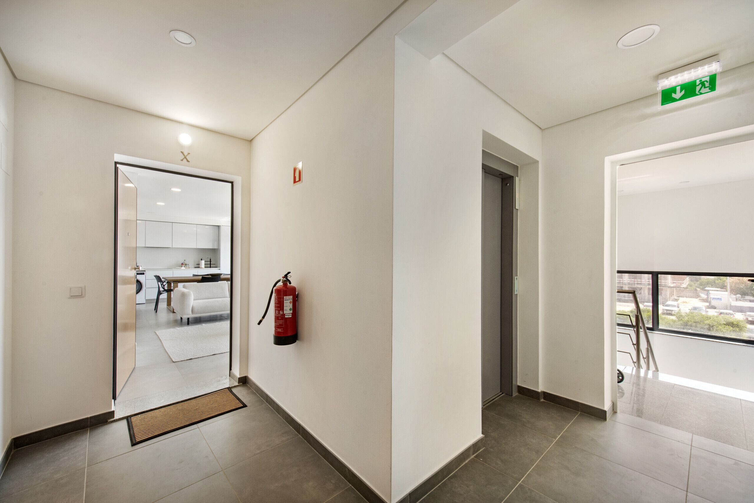 A hotel corridor with a fire extinguisher, a green emergency exit sign, and a view of the outside through a window.