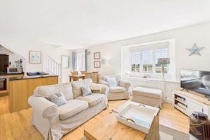 A modern living room with a sofa, armchairs, and a television. There is a kitchen area with a counter and appliances. A large window provides natural light and a view of the outdoors. - Host & Stay | Dove Cottage (Warkworth)