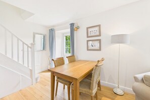 A dining area with a wooden table and wicker chairs, a floor lamp, and framed pictures on the wall. - Host & Stay | Dove Cottage (Warkworth)