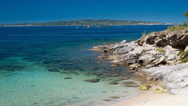 A coastal scene with clear waters, rocky shoreline, and a distant island with buildings.
