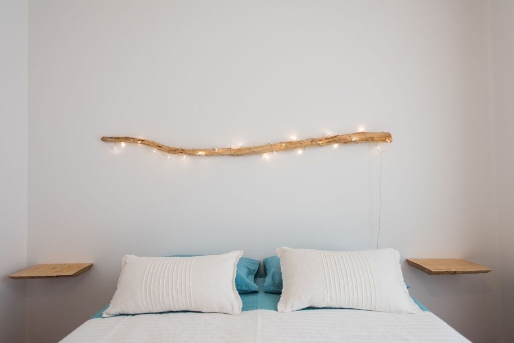 Cama com roupa de cama branca e azul, duas almofadas brancas e cabeceira de madeira com luzes de cordão.