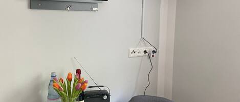 Una habitación con una televisión de pantalla plana montada en la pared, una mesa con una radio, una botella de agua y un florero con flores.