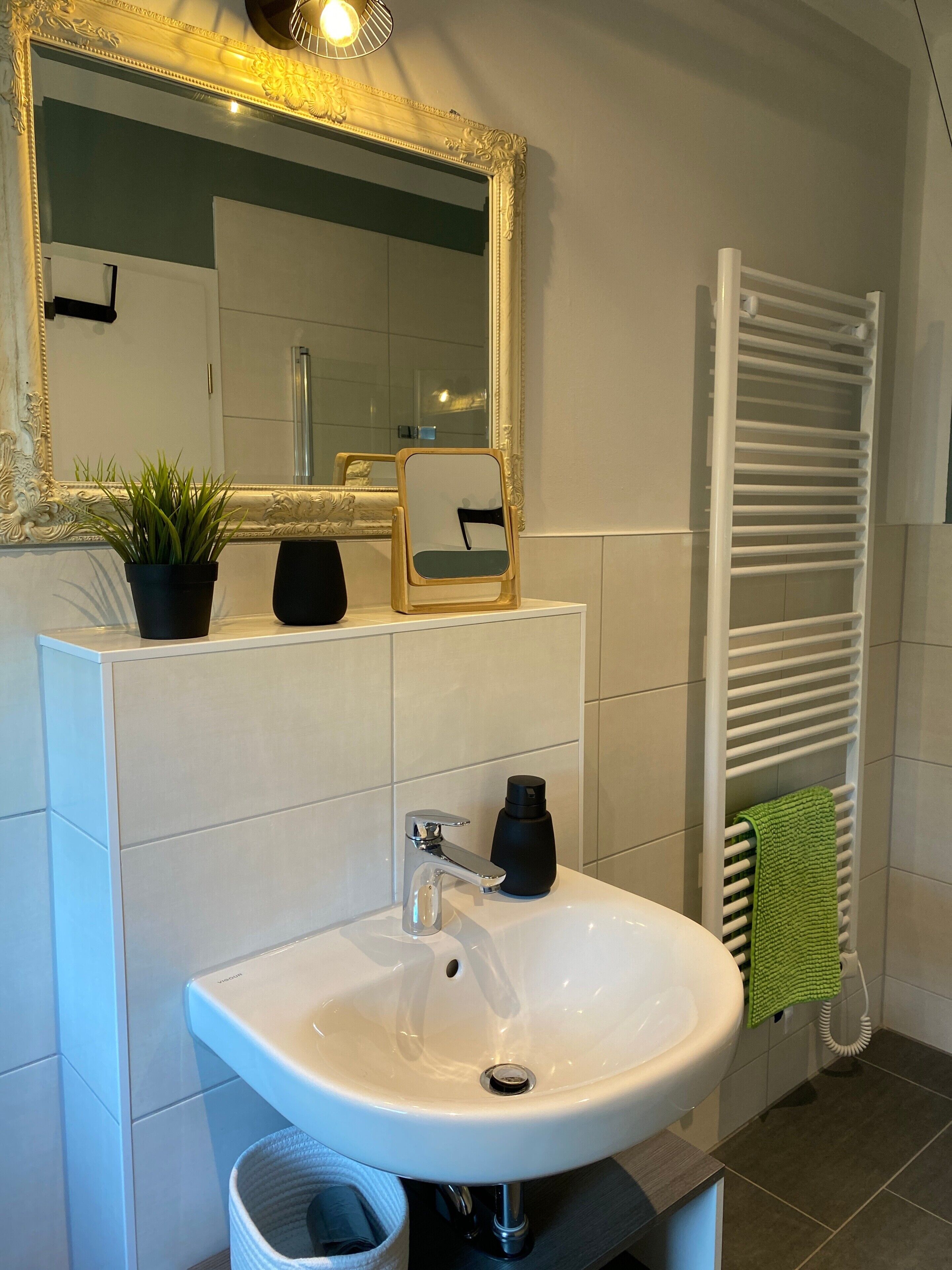 Une salle de bain avec un lavabo blanc, un miroir doté d’un cadre orné, un porte-serviettes et une plante en pot.