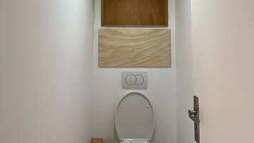 Une petite salle de bain comprenant des toilettes, une étagère en bois, un porte-rouleau de papier toilette et une poubelle.