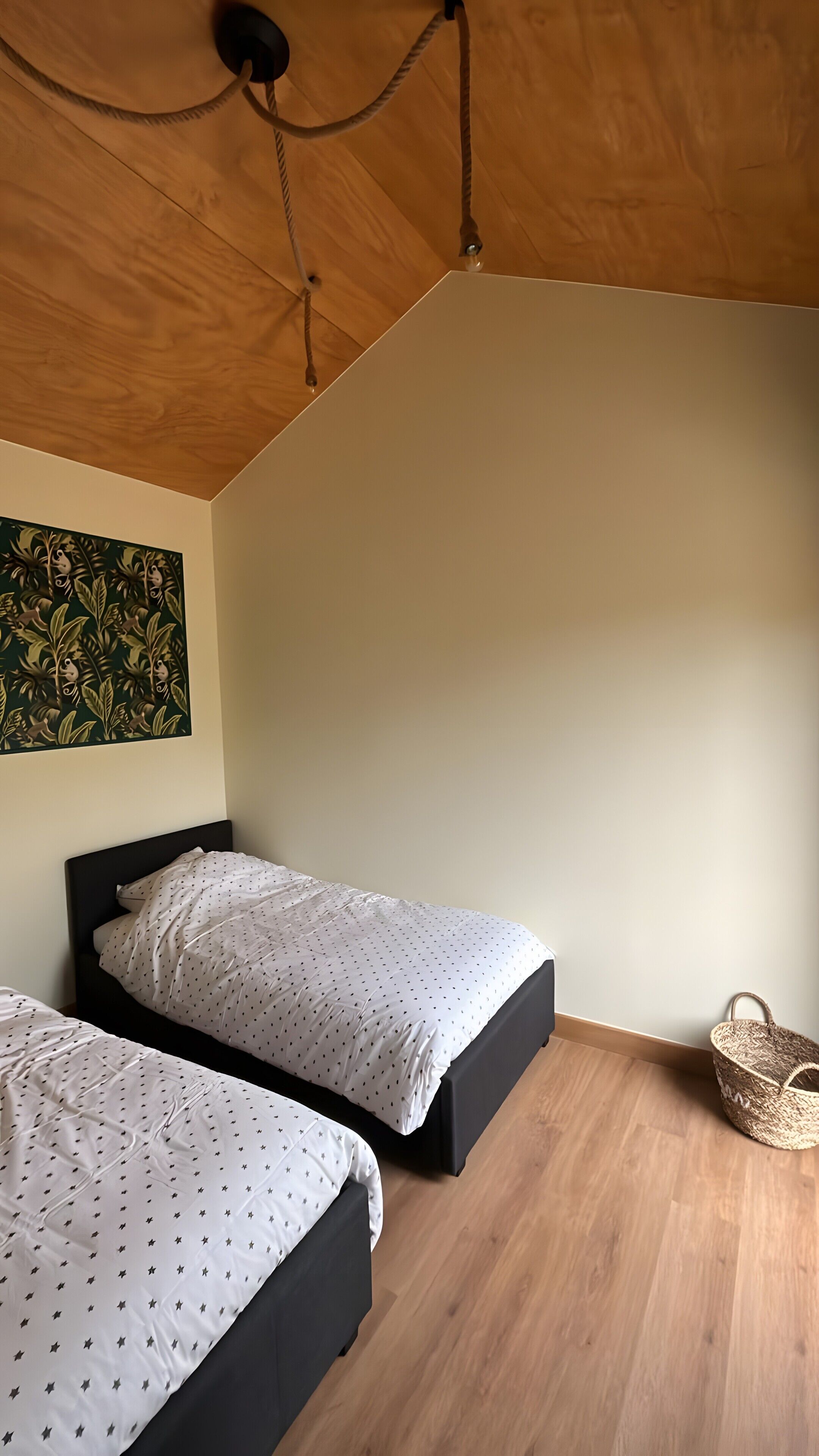 Una habitación con dos camas, techo de madera, pared con un diseño de hojas verdes y una cesta trenzada.