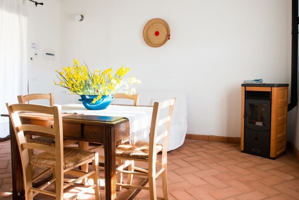 A dining area with a table, chairs, a vase of yellow flowers, a wall-mounted decorative plate, and a small heater.
