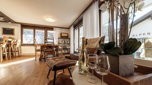 A modern living room with a wooden dining table, leather ottoman, and a large window with a view of a snowy landscape. - Apartment Sofi- Inspiringplaces Zermatt (Zermatt)