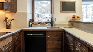 A kitchen with wooden cabinets, a dishwasher, a sink, and a window with a view of a snowy mountain. - Apartment Sofi- Inspiringplaces Zermatt (Zermatt)