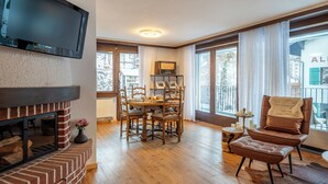 A living room with a fireplace, a television mounted on the wall, a dining table with chairs, and large windows with curtains. - Apartment Sofi- Inspiringplaces Zermatt (Zermatt)
