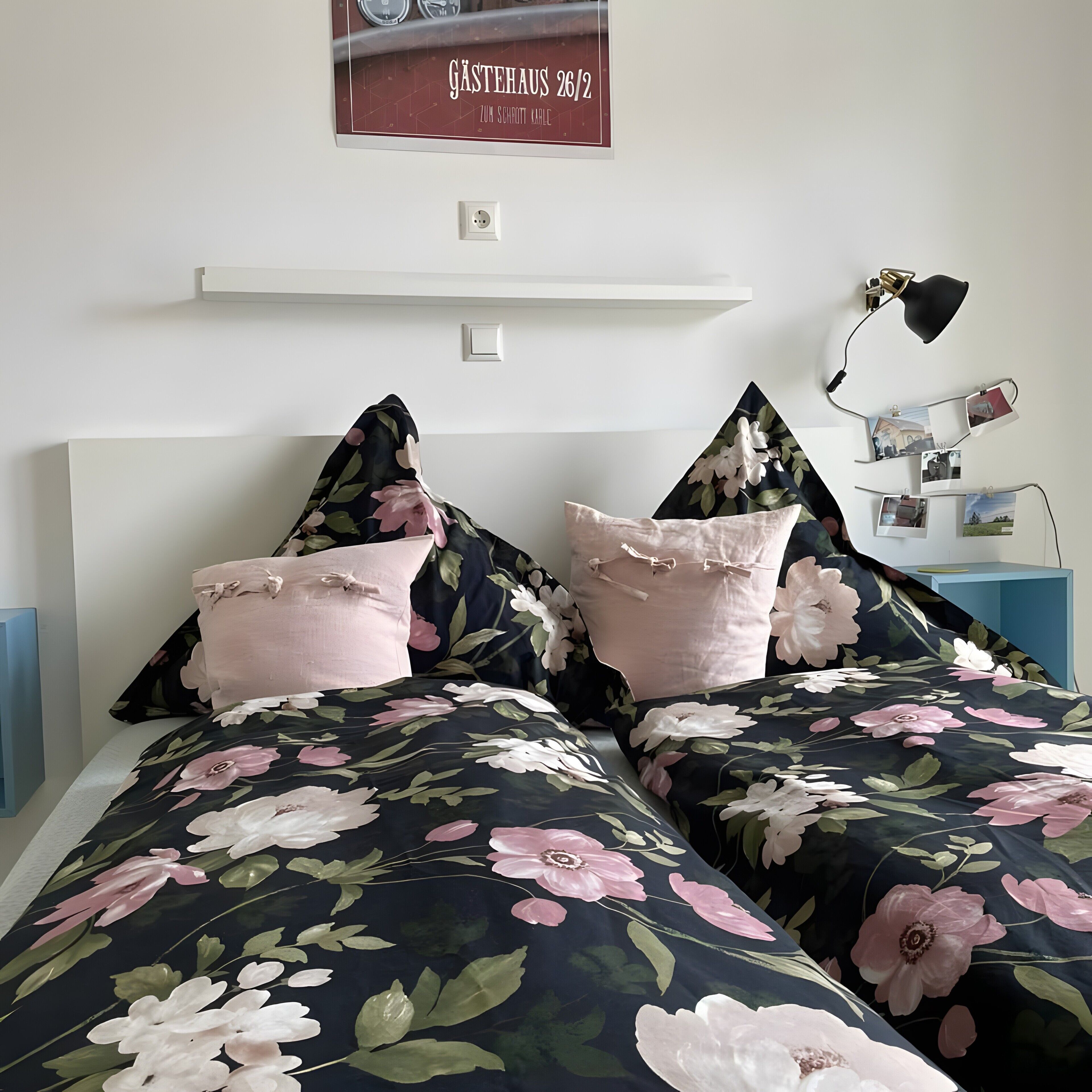 A bedroom with a bed featuring floral bedding, a wall-mounted shelf, and a desk with a lamp.