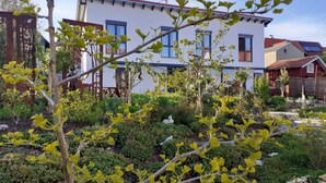 Zona residencial amb una casa blanca, un jardí amb plantes diverses i una tanca de fusta.