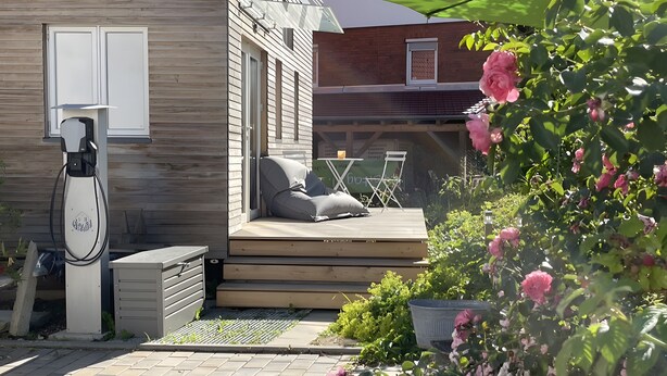 A wooden deck with a seating area, a charging station, and a garden with pink flowers. - Tiny Home 'Friedlinde' with Shared Terrace, Shared Garden and Wi-Fi (Bad Urach)