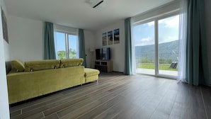A spacious living room with a yellow sofa, a flat-screen TV, and a balcony with a view of the mountains.
