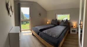 A bedroom with a large bed, a bedside table with a lamp, a window with a view of greenery, and a door leading to an outdoor area.