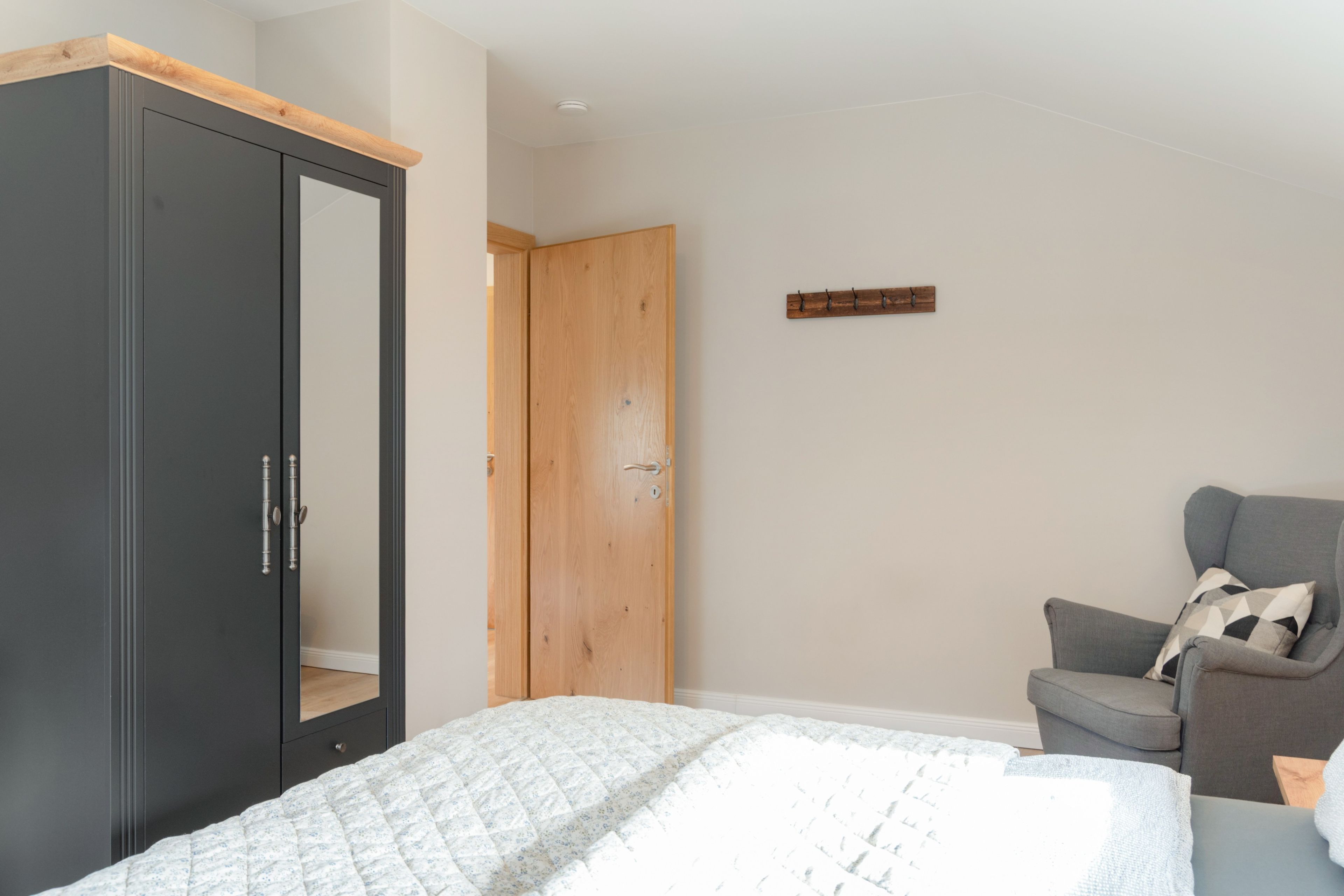 A bedroom with a bed, a wardrobe, a wooden door, a grey armchair, and a small wooden shelf.
