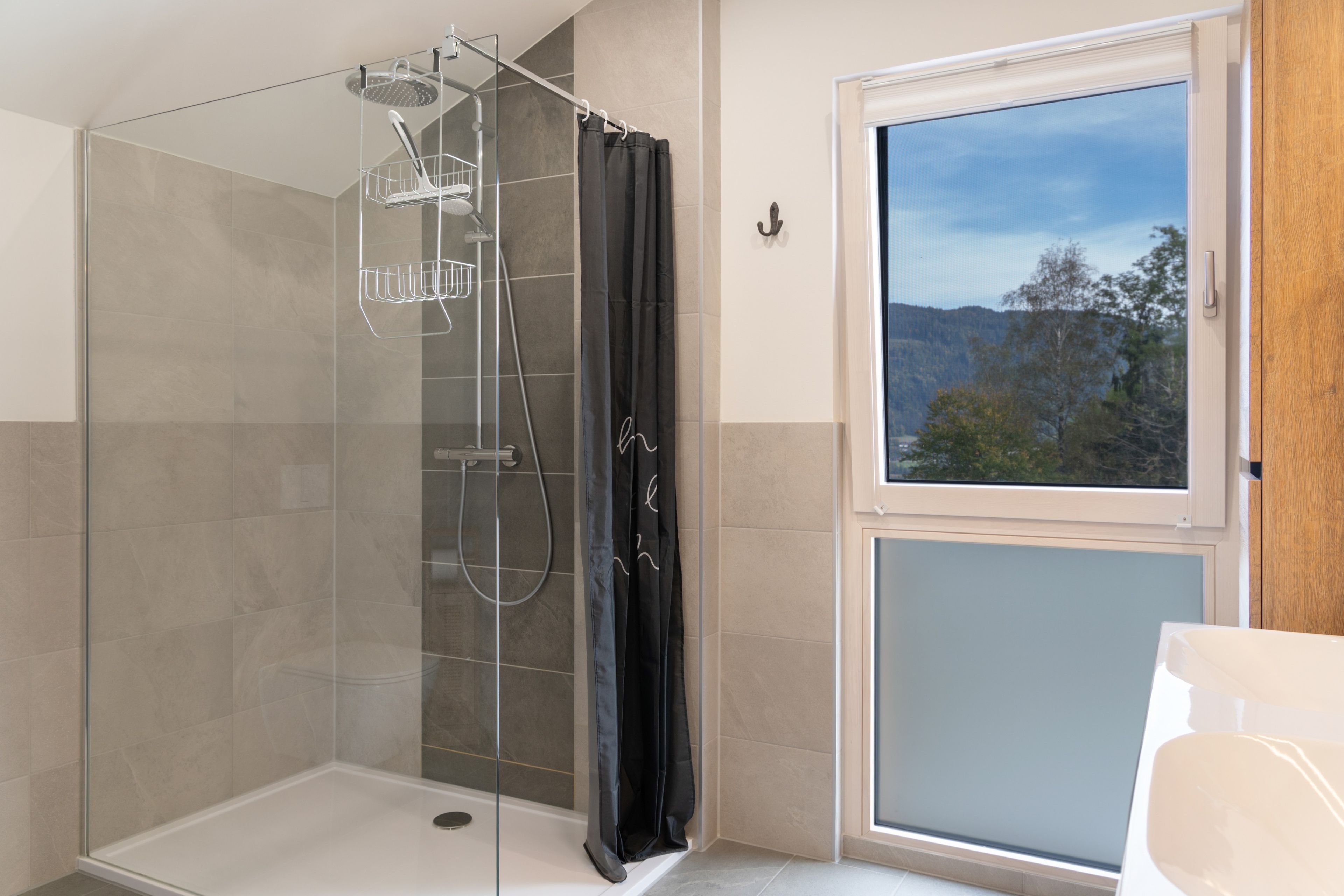 Ein modernes Badezimmer mit einer Duschkabine, einem Fenster mit Blick auf Bäume und einer weißen Badewanne.