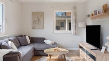 A modern living room with a grey sofa, a round wooden coffee table, a flat-screen TV, and a bookshelf with books and decorative items.