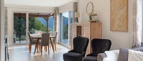 A spacious living area with a dining table, two armchairs, a sofa, and a large window offering a view of greenery.