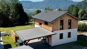 A modern house with a large deck, a covered patio, and a view of a lake and mountains. - Holiday Home 'Almrausch' with Lake View, Private Terrace and Wi-Fi (Bodensdorf)