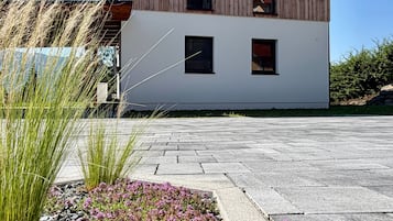 A modern two-story house with a wooden facade and white lower section, surrounded by a paved driveway and flowering plants.