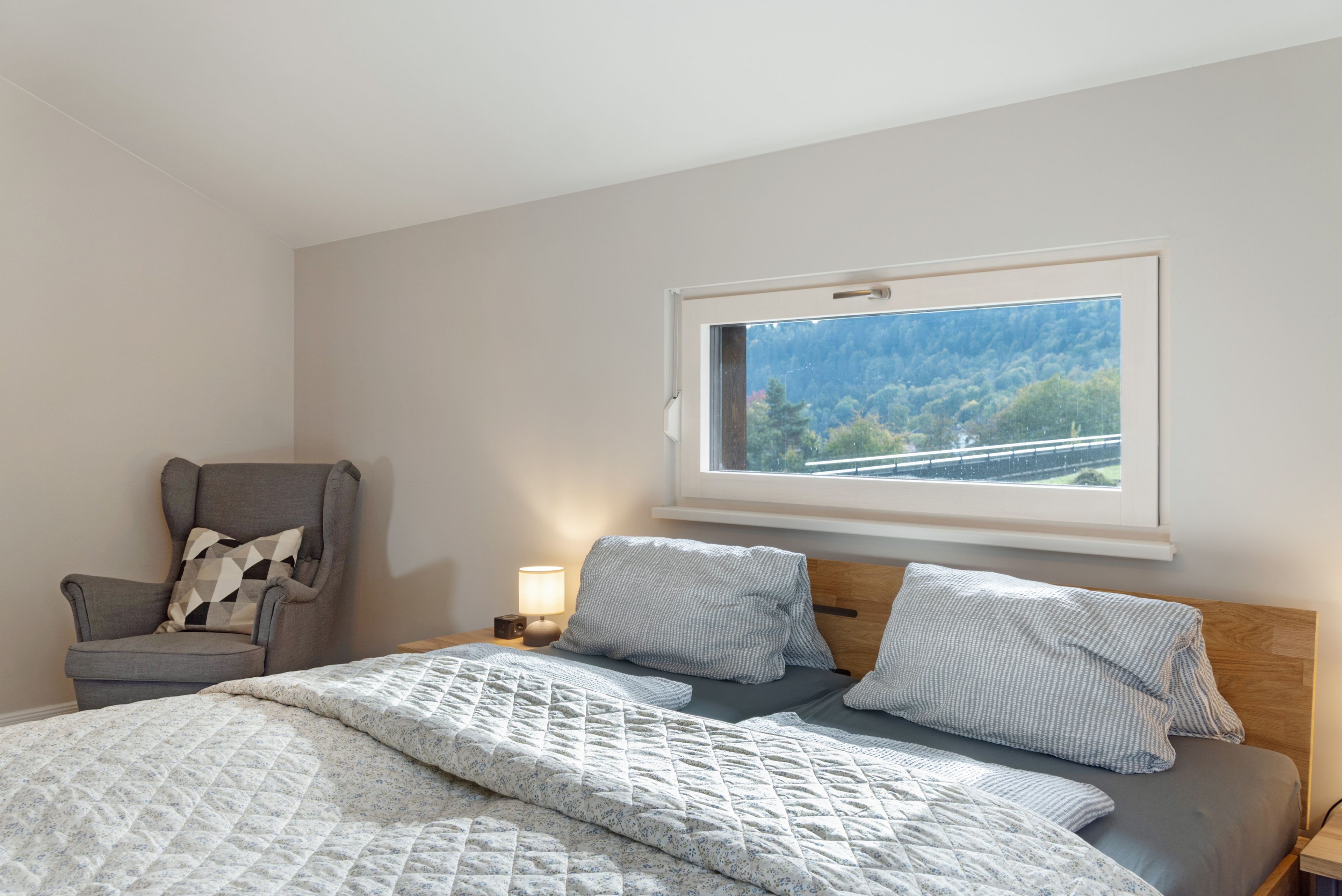 A bedroom with a bed, a chair, a bedside table with a lamp, and a window with a view of trees and a bridge.